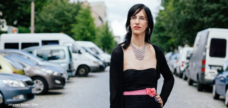 Frau mit Brille, roten Lippen, dunklen Haaren und schwarzem Minikleid steht mitten auf einer Straße, selbstbewusster Blick, links und rechts parkende Autos