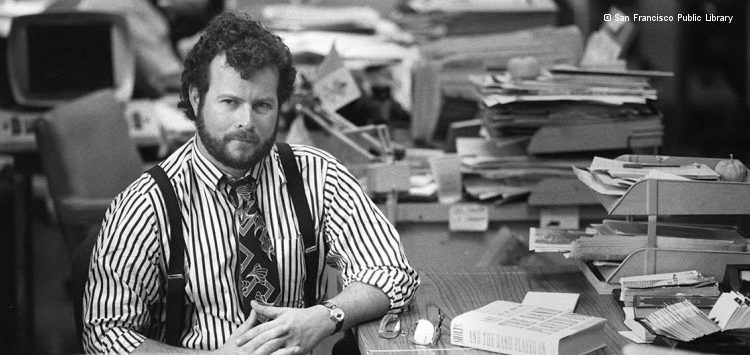 Schwarz-weiß-Foto von Randy Shilts: Mann mit Krawatte, Hemd und Hosenträgern sitzt an einem mit Büchern und Papier beladenen Schreibtisch