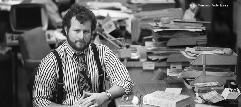 Schwarz-weiß-Foto von Randy Shilts: Mann mit Krawatte, Hemd und Hosenträgern sitzt an einem mit Büchern und Papier beladenen Schreibtisch