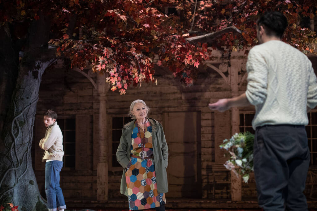 Beitragsbild zur Rezension des Theaterstücks Das Vermöchtnis; zwei Schauspieler und eine Schauspielerin unter einem Baum vor einem amerikanischen Holzhaus