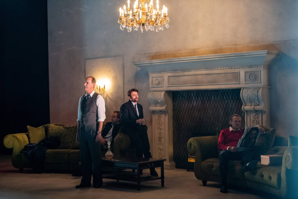 Beitragsbild zur Rezension des Theaterstücks Das Vermächtnis; Vier Schauspieler in einem herrschaftlichen Wohnzimmer mit zwei Sofas, großem Kamin und Kronleuchter