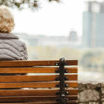 Altere Frau von hinten, sie sitzt auf einer Bank und blickt in die Ferne.