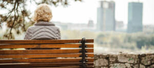 Altere Frau von hinten, sie sitzt auf einer Bank und blickt in die Ferne.