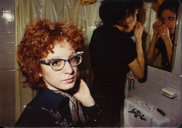 Älteres Selbstporträt Nan Goldins mit Brille und Halstuch, im Hintergund steht eine zweite feminine Person vor einem Toilettenspiegel
