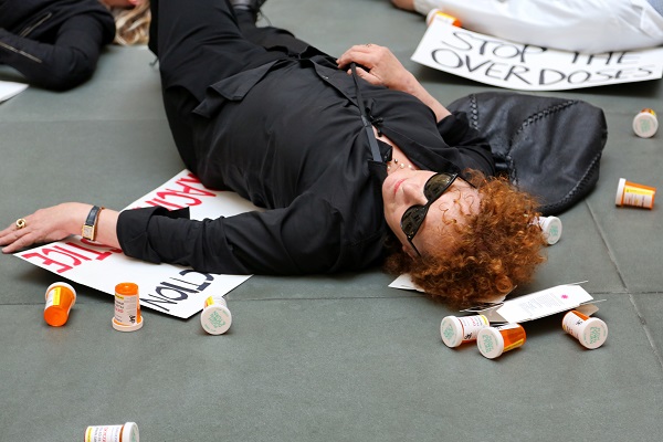 Nan Goldin liegt im Harvard Art Museum in schwarzer Kleidung mit Sonnenbrille auf dem Boden, unter ihr liegt ein nicht lesbares Protestschild, neben ihr zahlreiche leere Tablettendosen, im Hinterund ein Schild "Stop the Overdoses"