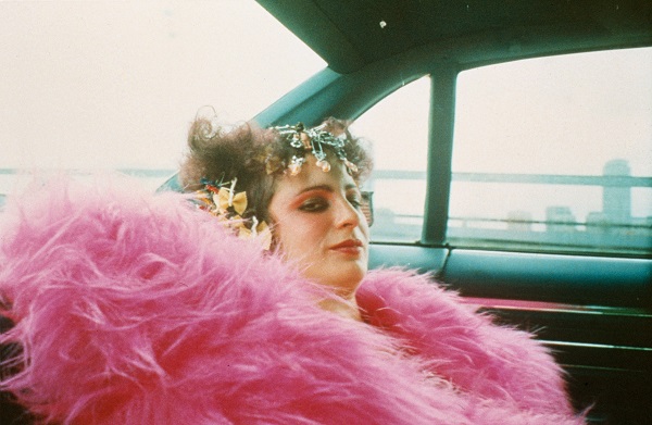 älteres Selbstporträt Nan Goldins mit pinker Federboa, buntem Make-Up und Kopfschmuck auf der Rückbank eines fahrenden Autos