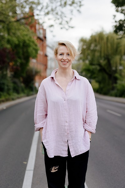 Porträt Kristina Shapran auf einer leeren Straße stehend, im lockeren rosa Leinenhemd und schwarzer Hose. 
