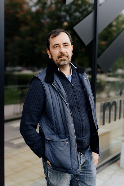 Porträt Volodymyr Zhovtyak, vor einer Fensterfrint stehend in Jeans, Strickjacke und Weste in dunkelblauen Farben