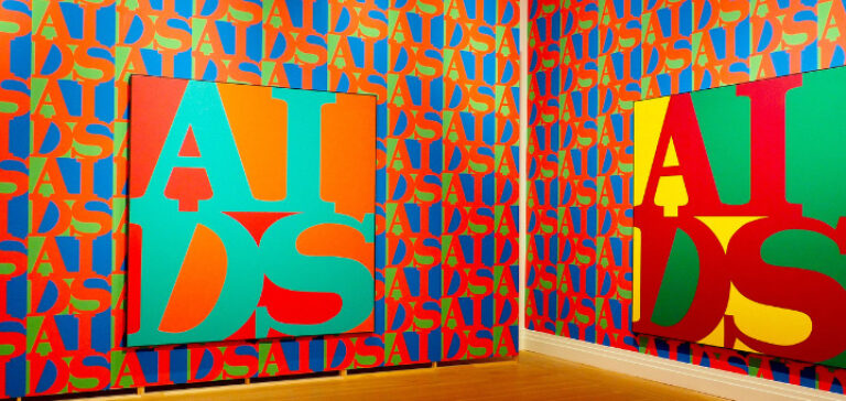 Blick in die Ausstellung: An zwei rot-blau-grün gemusterten Wänden hängt jeweils ein Gemälde mit dem AIDS-Logo in GRoßbuchstaben, einmal in Grün auf Rot, einmal in Rot auf gelb-grünem Hintergrund