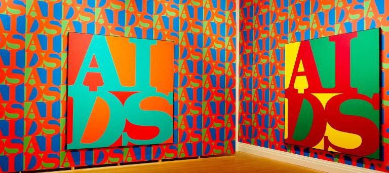 Blick in die Ausstellung: An zwei rot-blau-grün gemusterten Wänden hängt jeweils ein Gemälde mit dem AIDS-Logo in GRoßbuchstaben, einmal in Grün auf Rot, einmal in Rot auf gelb-grünem Hintergrund
