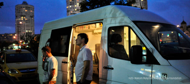 Zwei männliche Personen stehen vor der offenen Seitentür eines beleuchteten Kleinbusses in einer nächtlichen Stadtszene. Im Hintergrund Hochhäuser.