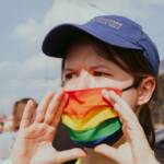 Demoszene: Person mit Regenbogenmaslke uns Basecap hält rufend die Hände vor dem Mund