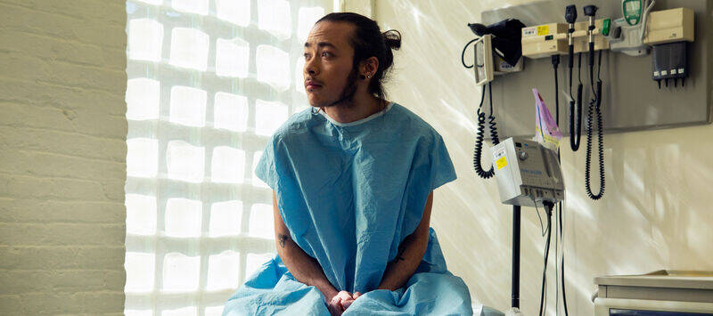 A genderqueer person sitting in a hospital gown sitting in an exam room