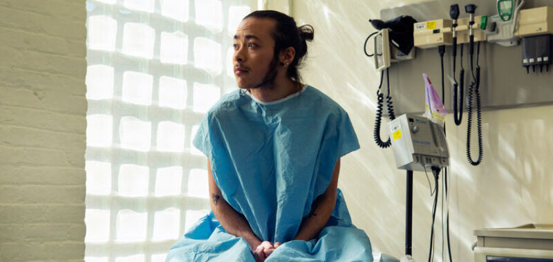 A genderqueer person sitting in a hospital gown sitting in an exam room