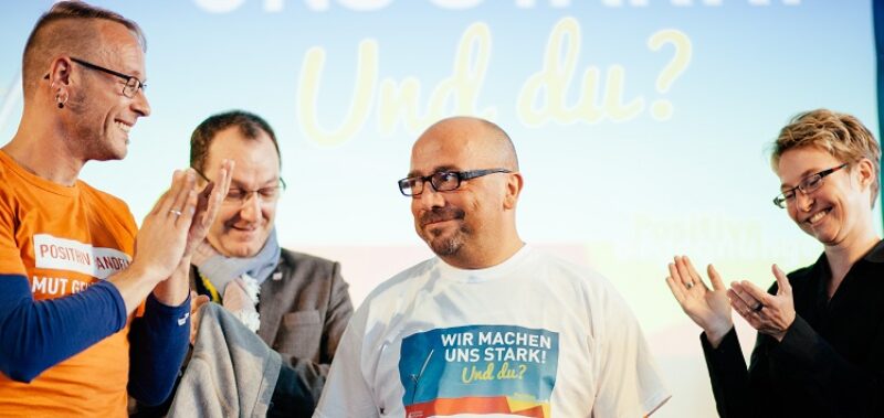 Drei Personen applaudieren einer Person im weißen T-Shirt mit der Aufschrift