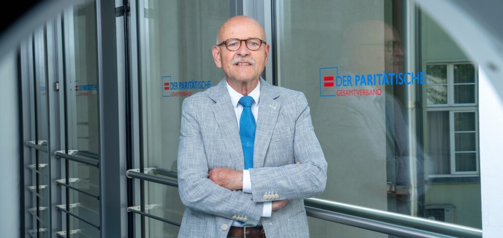Porträt Rolf Rosenbrock vor Glastür mit dem Logo des Paritätischen Wohlfahrtsverbands