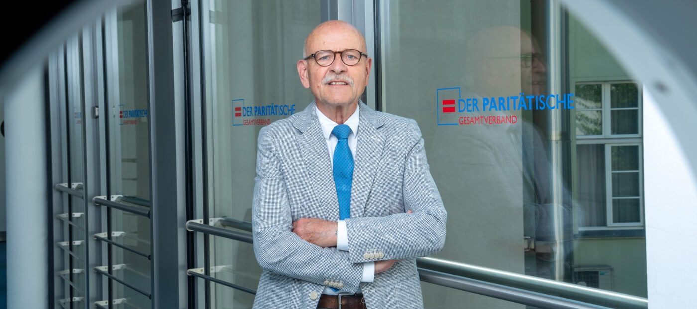 Porträt Rolf Rosenbrock vor Glastür mit dem Logo des Paritätischen Wohlfahrtsverbands