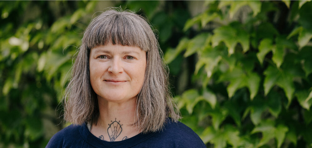 Porträtfoto weibliche Person mit halblangen Haaren und blauem Oberteil, iim Hintergrund grüne Pflanzen