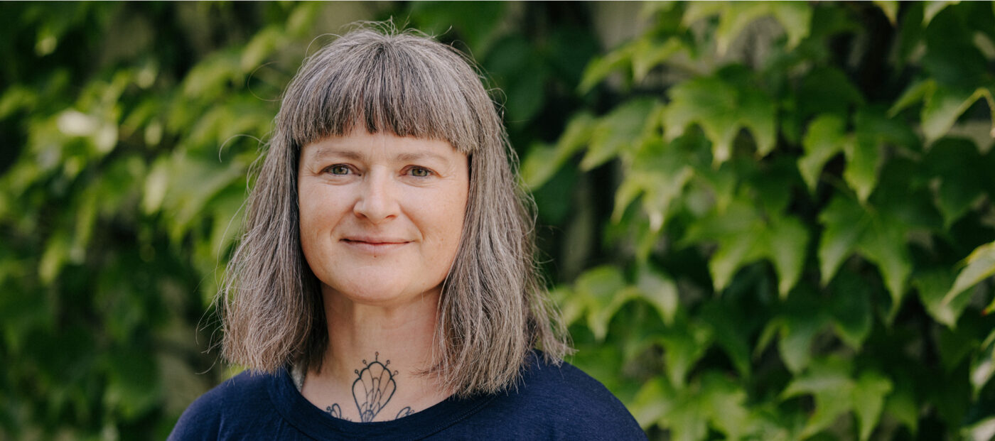 Porträtfoto weibliche Person mit halblangen Haaren und blauem Oberteil, iim Hintergrund grüne Pflanzen