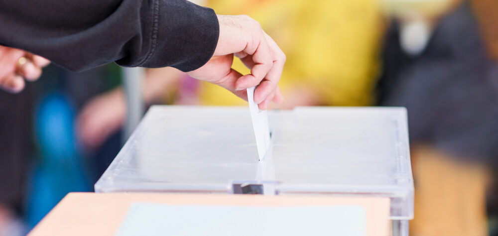 Eine Person, zu sehen ist nur der Unterarm mit der Hand, steckt einen Stimmzettel in eine Wahlurne