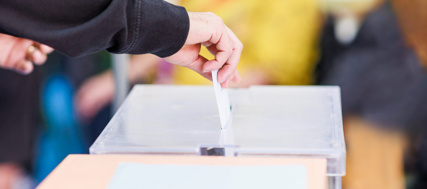 Eine Person, zu sehen ist nur der Unterarm mit der Hand, steckt einen Stimmzettel in eine Wahlurne