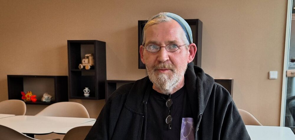 Porträt Stephan Raabe, im Wartezimmer sitzend mit schwarzer Jacke, Brille, grauer Mütze