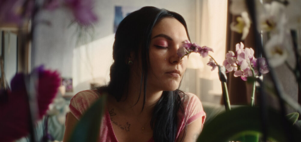 Szene aus dem Film „Vena“: Jenny (gespielt von Emma Nova) mit pinkem Lidschatten und pinkem Shirt sowie langen schwarzen Haaren riecht mit geschlossenen Augen an einer Orchidee.