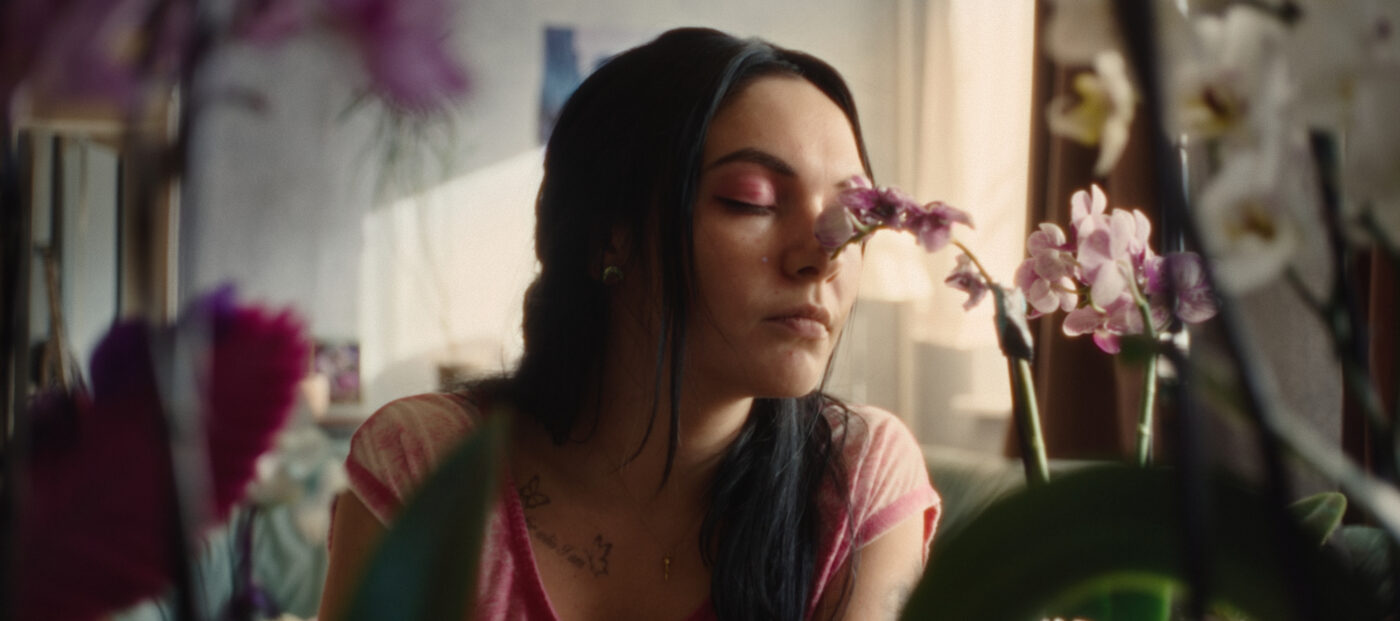 Szene aus dem Film „Vena“: Jenny (gespielt von Emma Nova) mit pinkem Lidschatten und pinkem Shirt sowie langen schwarzen Haaren riecht mit geschlossenen Augen an einer Orchidee.