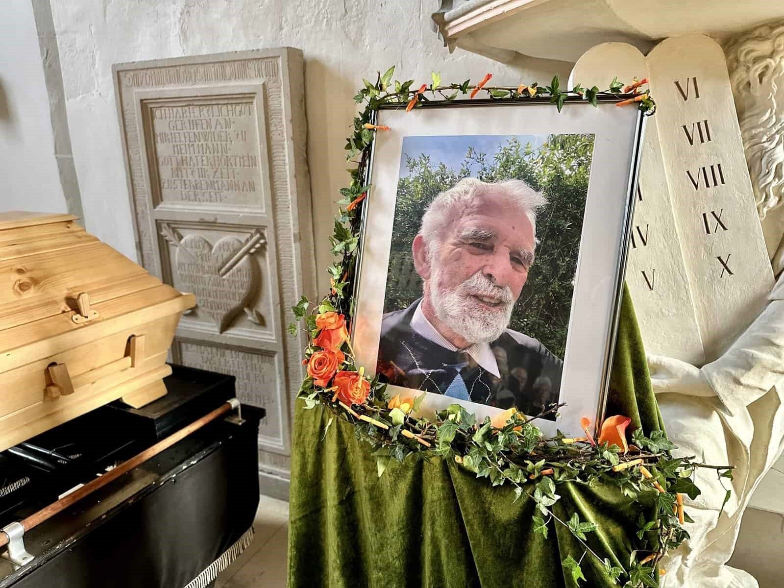 Szene der Beisetzung: ein Holzsarg und ein gerahmtes Porträt von Petrus Ceelen mit Rosengesteck