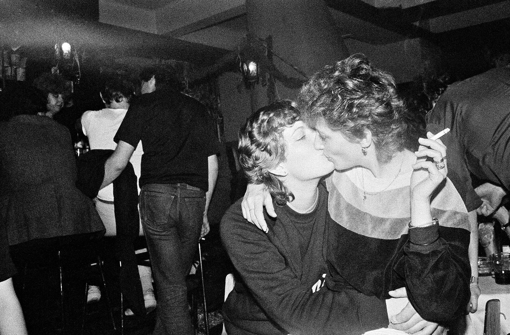 schwarz-weiß Foto einer Clubszene: zwei küssende Frauen, im Hintergrund Menschen an der Bar