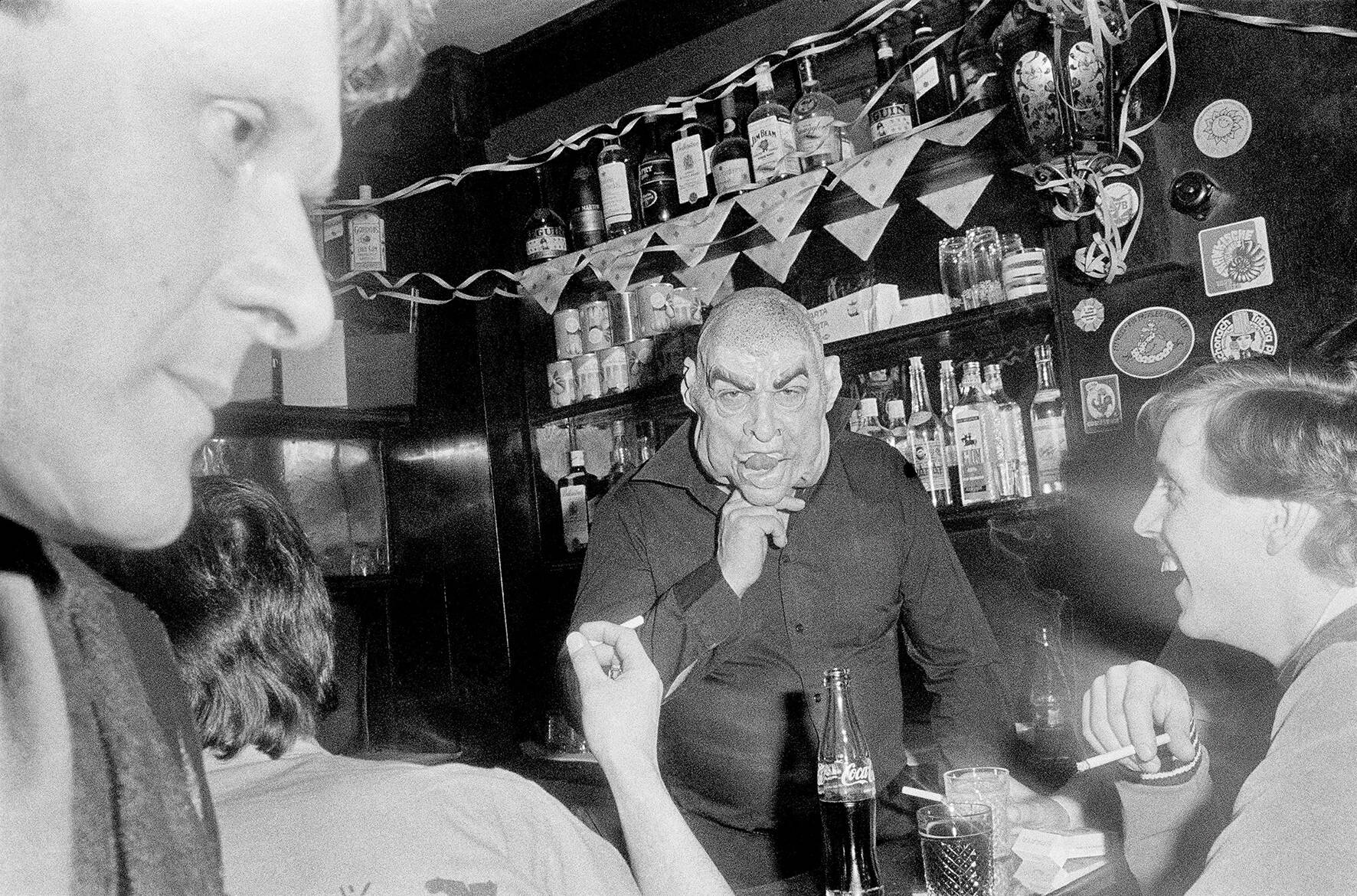 männliche Person mit Monstermaske steht hinter einer Bar, davor drei unscharf zu erkennende Personen