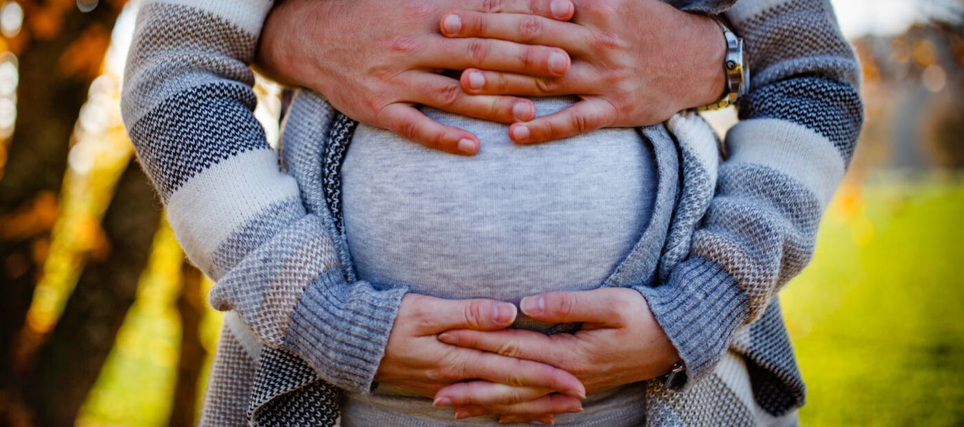 Eine schwangere Person umfasst den Bauch mit ihren Händen, von hinten wir sie von einer anderen Person umfasst.