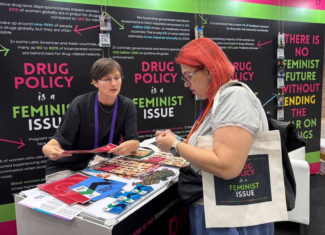 zwei Personen sprechen an einem Messestand, auf dem bunte Flyer liegen, dahinter Plakate mit der Auschrift