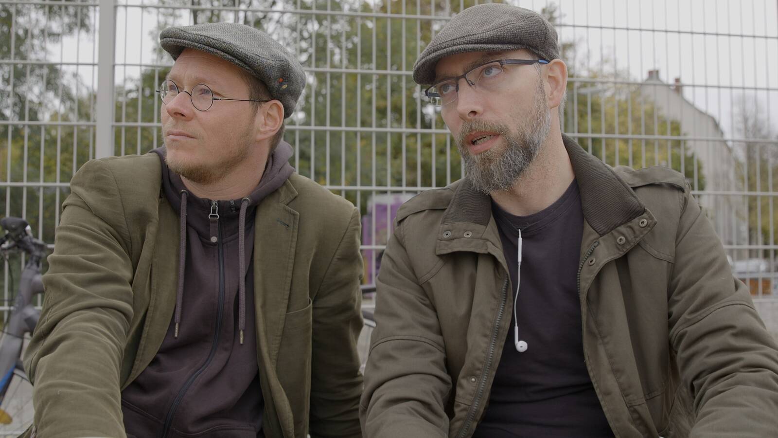 Zwei Männer mit Mütze und dunkler KLeidung sitzen nebeneinander vor einem Zaun und blicken in die Ferne