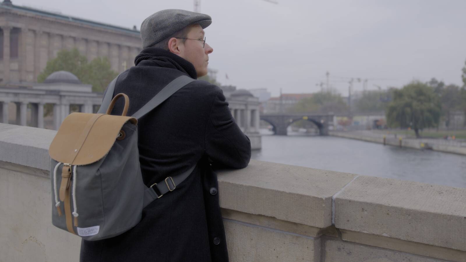 Person mit Rucksack und Mütze steht auf einer Brücke und blickt über den Fluss