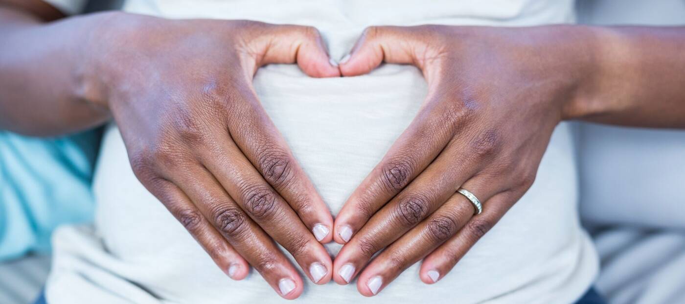 Symbolbild zu Rassismus in der Geburtshilfe: Schwangere Schwarze Person im weißen T-Shirt formt mit ihren Händen ein Herz auf dem Bauch
