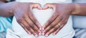 Symbolbild zu Rassismus in der Geburtshilfe: Schwangere Schwarze Person im weißen T-Shirt formt mit ihren Händen ein Herz auf dem Bauch