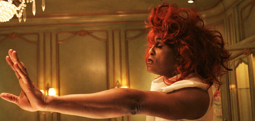 Foto einer Person mit roter Perrücke und nach vorn gestreckten Armen in einem grün-gold gestrichenen Raum