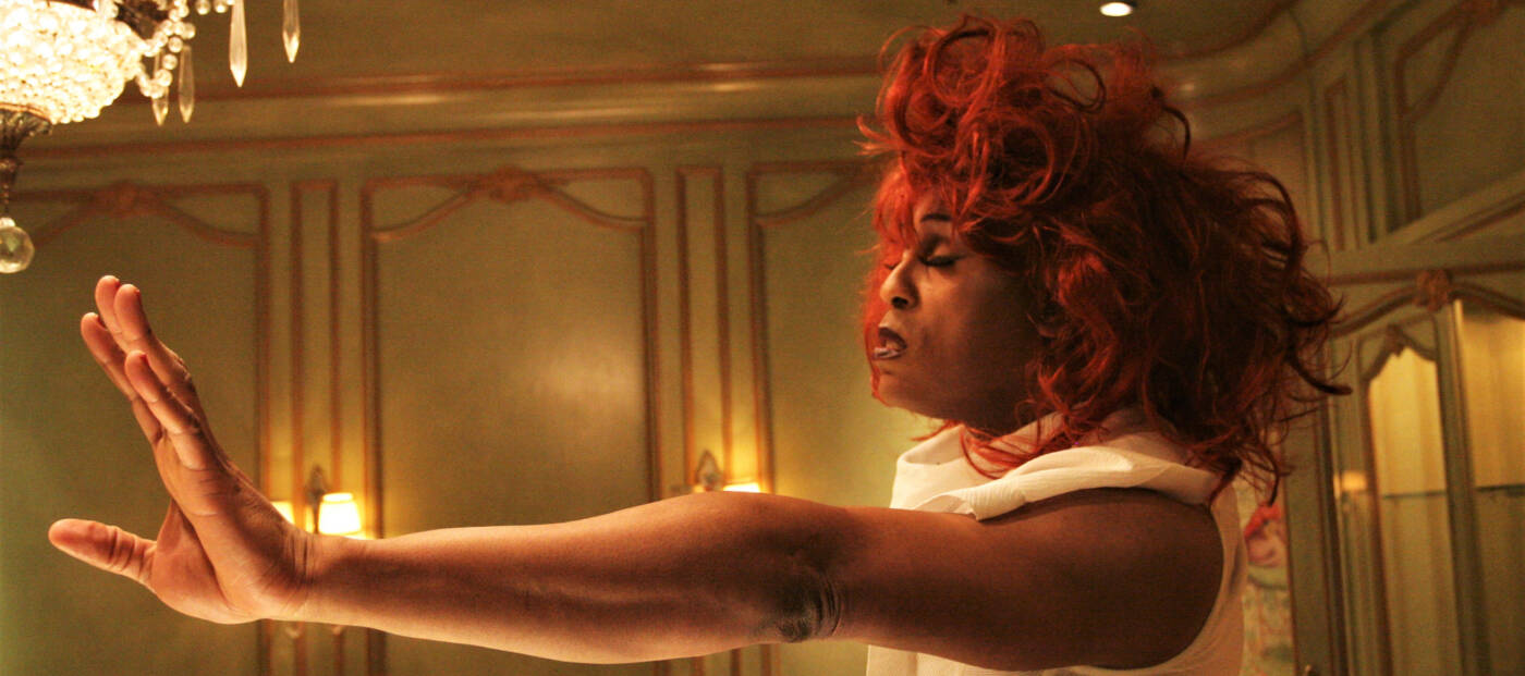 Foto einer Person mit roter Perrücke und nach vorn gestreckten Armen in einem grün-gold gestrichenen Raum