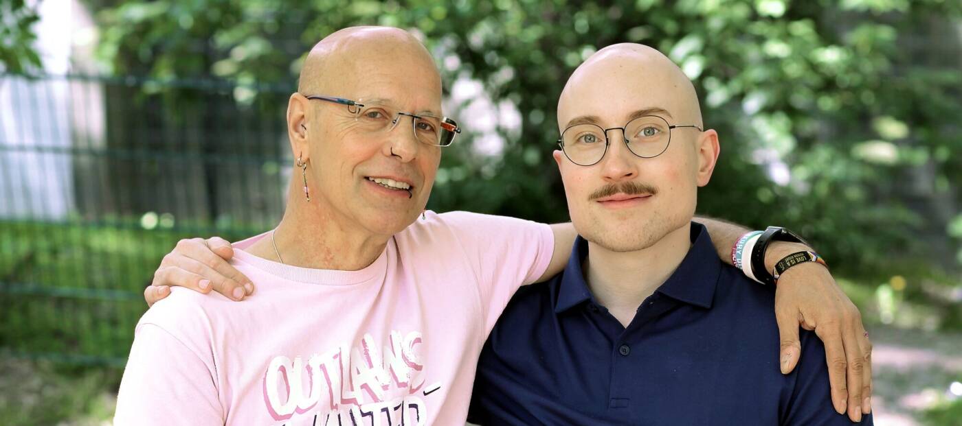 Die beiden Interviewten stehen Arm in Arm in einem sonnigen Park.