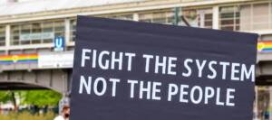 Bild zum Beitrag "Aidshilfe erinnert daran: Nicht die Menschen sind falsch, das System ist es oft"; im Bild eine schwarzes Plakat mit dem Text "Fight the system, not the people"