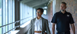 Zwei Personen gehen durch einen Flur, eine trägt ein Namensschild mit "Justiz", die andere Akten unterm Arm