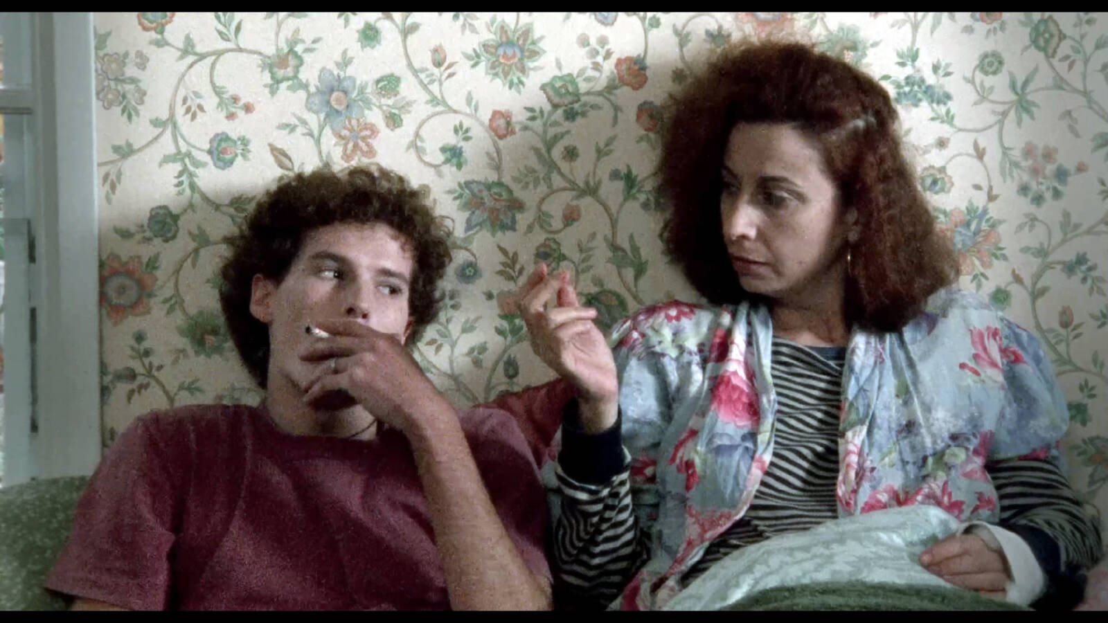 Zwei Personen sitzen rauchend nebeneinander vor einer geblümten Tapete