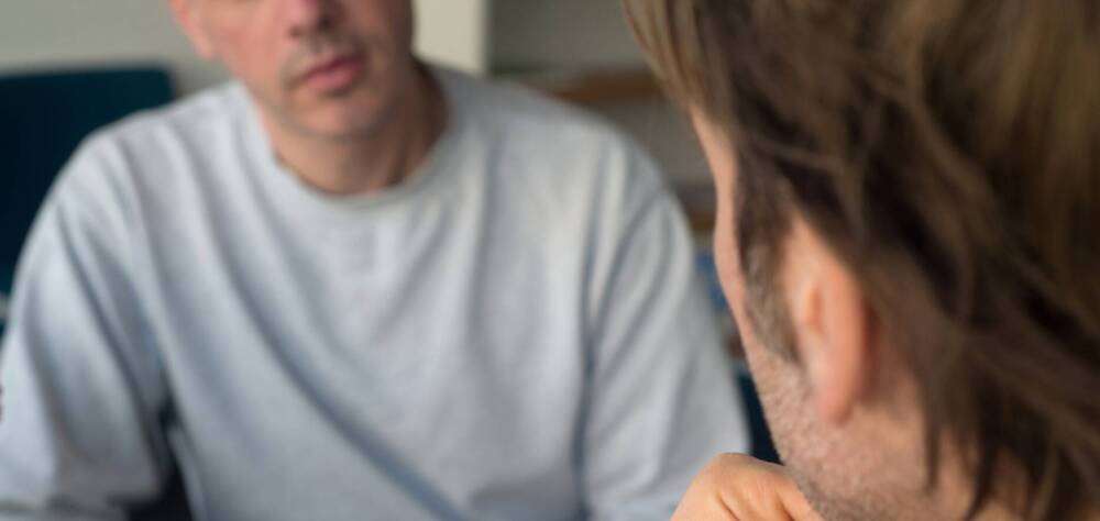 Zwei männlich gelesene Personen sitzen sich in einem ernsten Gespräch gegenüber.