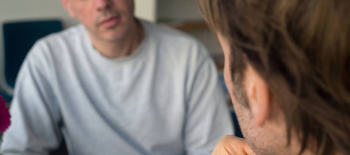 Zwei männlich gelesene Personen sitzen sich in einem ernsten Gespräch gegenüber.