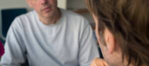 Zwei männlich gelesene Personen sitzen sich in einem ernsten Gespräch gegenüber.