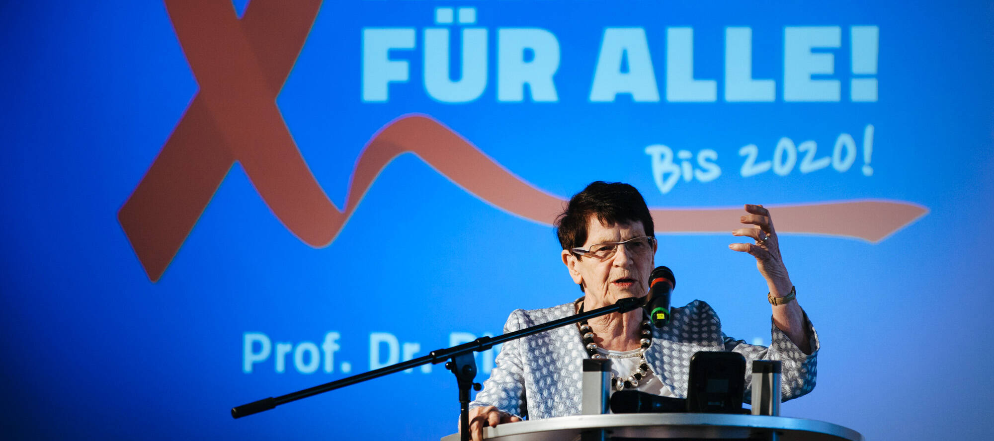 Rita Süssmuth am Redepult, Hintergrund: eine rote Aidsschleife und die Schrift 