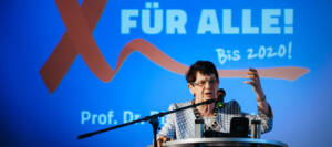 Rita Süssmuth am Redepult, Hintergrund: eine rote Aidsschleife und die Schrift "Kein Aids für alle!"