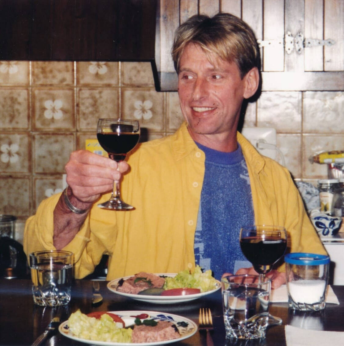 Lächernder Mann im gelben Hemd erhebt ein Glas Rotwein am Esstisch